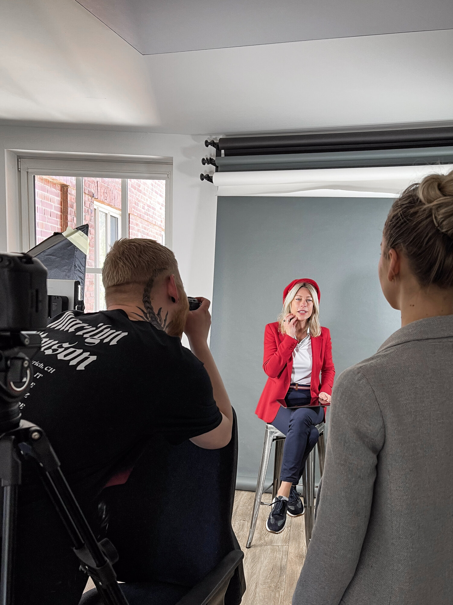 Fotoshooting: Frau mit rotem Blazer und Hut sitzt auf einem Hocker vor grauem Hintergrund